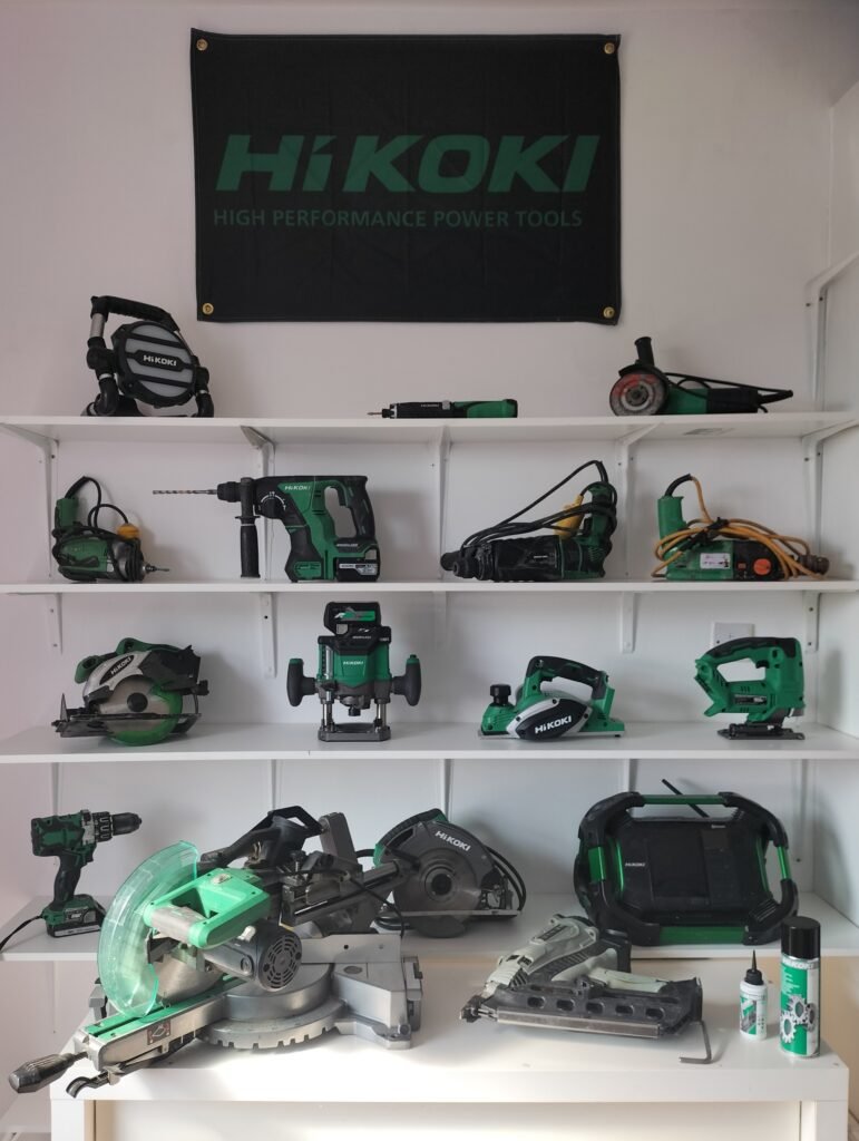 Hikoki power tools display: A collection of various Hikoki power tools upon several shelves. A Hikoki banner is at the top of the display which comprises both mains and cordless battery tools by Hikoki. In the foreground there is a 110v Hikoki C8FSHG sliding compound mitre saw along with a Hitachi Koki NR90GC2 90mm Nailgun + Cleaning kit. On the shelves above there are Hikoki power tools which (from top to bottom) consist of  (top shelf left to right) the Hikoki UB18DGLL07 18V Worklight, Hikoki 3DL2/Jlz cordless screwdriver, Hikoki G12STX 110V Angle grinder (2nd from top shelf left to right) Hitachi W8VB2 110v Tek Gun, Hikoki Brushless DH18DBL Rotary Hammer Drill, Hitachi 110v DH26PX Rotary Hammer drill, Hitachi Koki DV20T 110v Hammer Drill, (3rd from top shelf left to right) Hikoki C18DSL 18V cordless circular saw, Hikoki brushless multivolt Router M3612DA, Hikoki P18DSL cordless Planer, Hikoki CJ 18DA cordless Jigsaw 135mm 18v, (bottom shelf left to right) Hikoki cordless combi drill DV 18DBXL, Hikoki C7SB3 185mm Circular saw 110v, Hikoki UR18DSDL cordless radio Bluetooth FM DAB