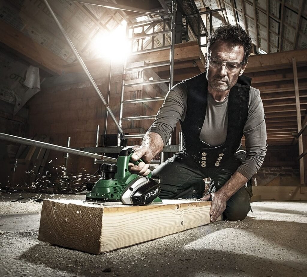 A carpenter operates the Hikoki P18DSL cordless Planer in an internal construction project. He is wearing safety glasses while planing a length of timber which is resting on the ground.